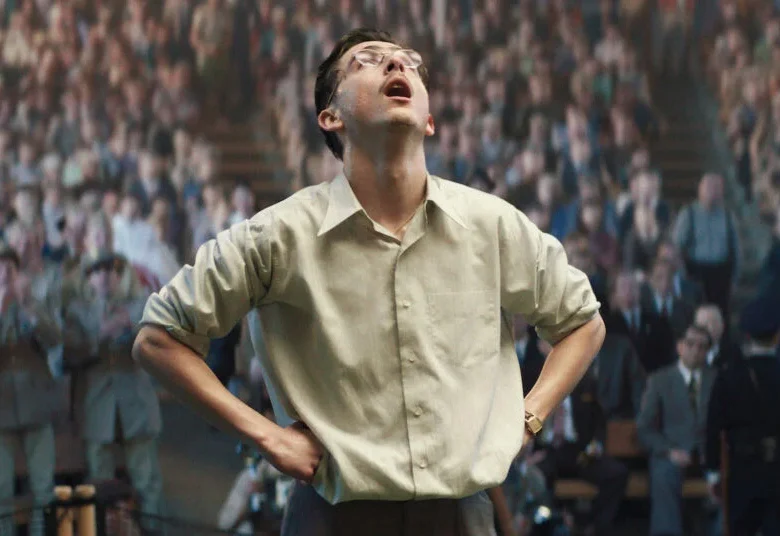 Na pierwszym planie stoi młody mężczyzna z wyrazem zmęczenia i ulgi na twarzy. W tle na trybunach stoją patrzący na niego ludzie. Kilka osób klaszcze.