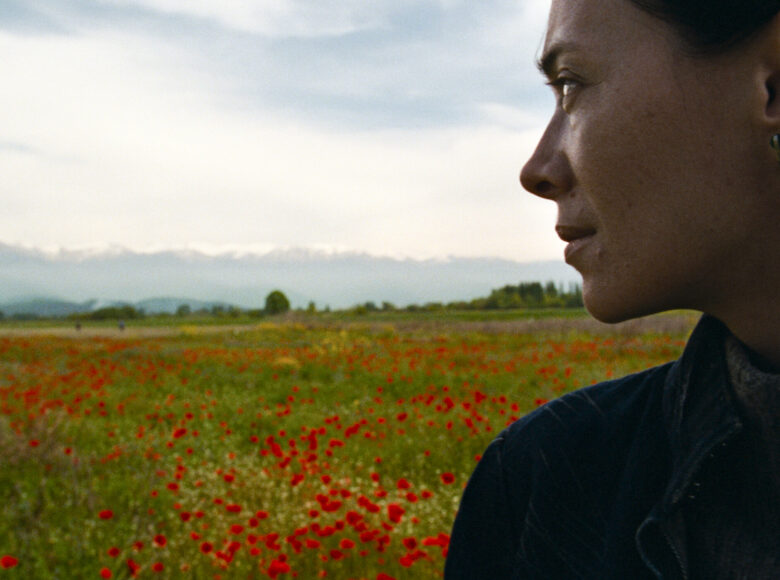 Zbliżenie na twarz kobiety, odwróconą do nas profilem. Kobieta patrzy przez ramię na łąkę pełną kwiatów, która znajduje się za jej plecami. Nad łąką rozpościera się jasne niebo.