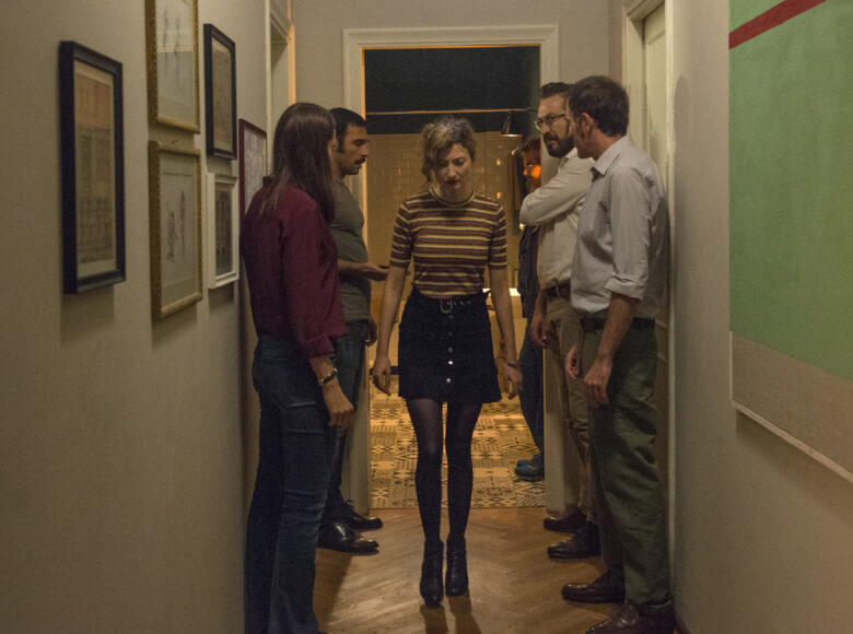 Wąski przedpokój w mieszkaniu. Pod przeciwległymi ścianami stoją naprzeciwko siebie cztery osoby - kobieta i trzech mężczyzn. Między nimi przechodzi kobieta wychodząca z pokoju, który znajduje się na końcu przedpokoju.