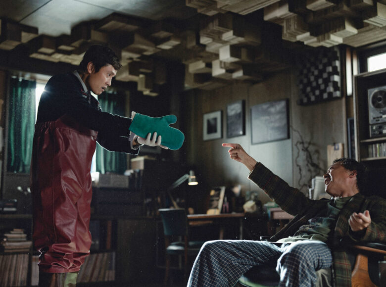 W pokoju dwóch azjatyckich mężczyzn. Starszy siedzi na fotelu. Młodszy stoi naprzeciwko starszego i wyciąga w jego stronę dłoń odzianą w rękawicę kuchenną. Za mężczyznami widzimy półki z książkami, biurko, wiszące nad biurkiem obrazki oraz półkę z magnetofonem szpulowym.
