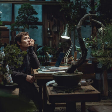 Pokój pełen roślin doniczkowych. Przy stole siedzi chłopak i podpiera ręką twarz, patrząc przed siebie w zamyśleniu. Na stole leżą kartki i stoi zapalona lampka do pracy. Z tyłu widzimy duże okno, a przez nie ciemne, nocne niebo. W pokoju panuje półmrok.