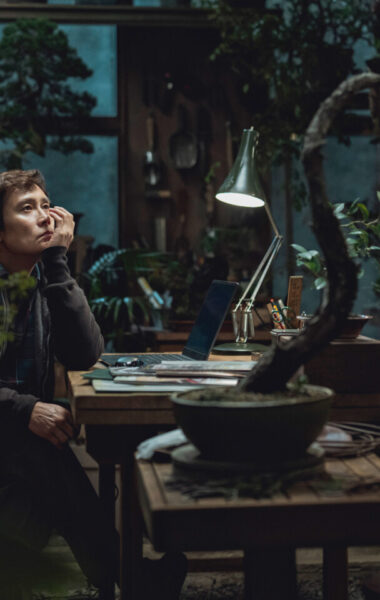 Pokój pełen roślin doniczkowych. Przy stole siedzi chłopak i podpiera ręką twarz, patrząc przed siebie w zamyśleniu. Na stole leżą kartki i stoi zapalona lampka do pracy. Z tyłu widzimy duże okno, a przez nie ciemne, nocne niebo. W pokoju panuje półmrok.