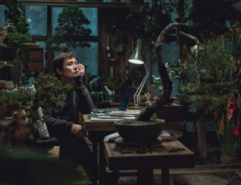 Pokój pełen roślin doniczkowych. Przy stole siedzi chłopak i podpiera ręką twarz, patrząc przed siebie w zamyśleniu. Na stole leżą kartki i stoi zapalona lampka do pracy. Z tyłu widzimy duże okno, a przez nie ciemne, nocne niebo. W pokoju panuje półmrok.