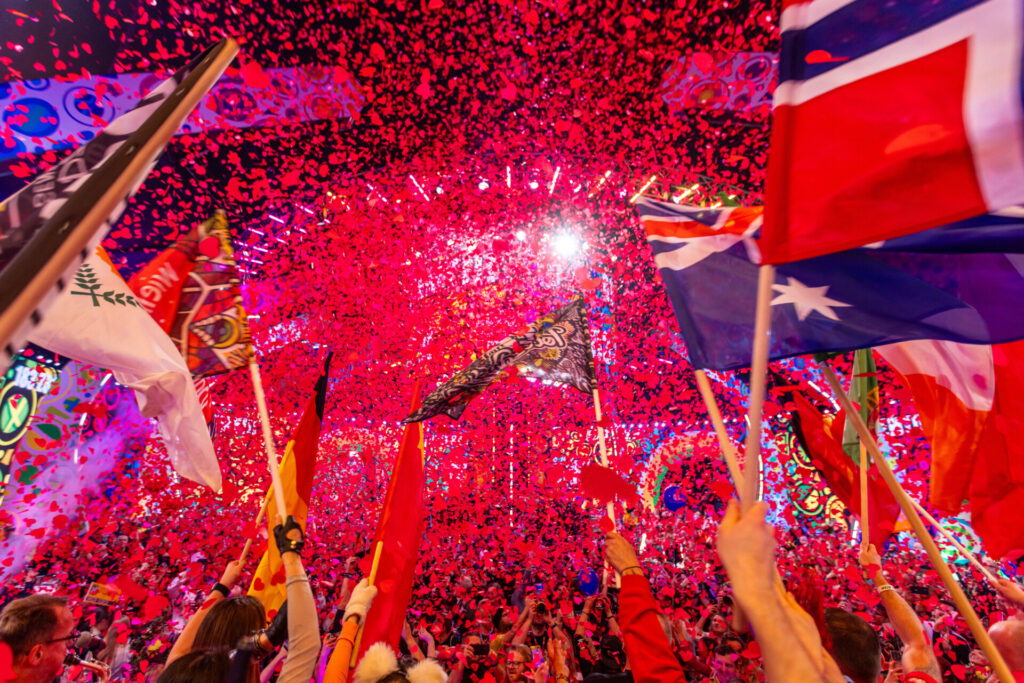 Kolorowa fotografia, utrzymana w klimacie agresywnej, świecącej czerwieni, przedstawia napierwszym planie przedramiona wielu osób trzymających różnokolorowe chorągiewki. Drugi plan tworzy wielkie skupisko ludzi uczestniczących w masowej imprezie. Zdjęcie ma nierealistyczny charakter. Rozciągający się w tle tłum pozostaje nieostry. Oświetlony czerwonym światłem przypomina gęsty, rozżarzony kosmos – mgławicę złożoną z tysięcy świecących postaci.