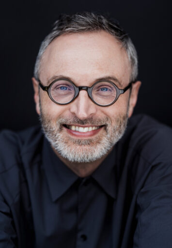 Zbliżenie na twarz mężczyzny w średnim wieku. Mężczyzna lekko się uśmiecha. Ma siwiejące włosy i brodę. Nosi okulary i granatową koszulę.