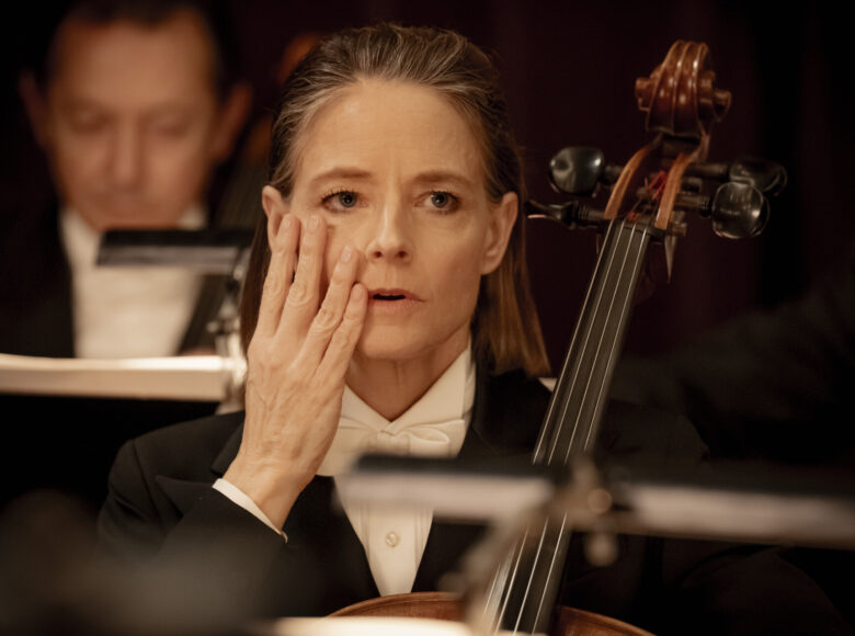 Zbliżenie na twarz i ramiona kobiety ubranej w czarną marynarkę i biała koszulę z białą muchą. Kobieta jedną ręką trzyma przed sobą wiolonczelę. Drugą rękę przykłada do policzka. Ma zaniepokojony wyraz twarzy.