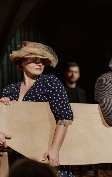 Przedstawienie teatralne. Na scenie kobieta i dwóch mężczyzn w staroświeckich strojach. Jeden z mężczyzn ma wioślarską czapkę na głowie. Wszyscy troje trzymają płaską sklejkę, która jest wycięta w kształt kojarzący się z kajakiem.