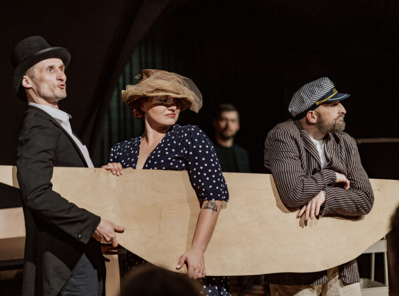 Przedstawienie teatralne. Na scenie kobieta i dwóch mężczyzn w staroświeckich strojach. Jeden z mężczyzn ma wioślarską czapkę na głowie. Wszyscy troje trzymają płaską sklejkę, która jest wycięta w kształt kojarzący się z kajakiem.