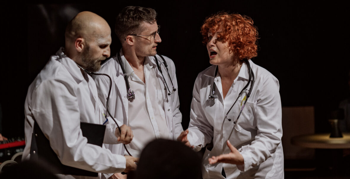 Przedstawienie teatralne. Na scenie stoją obok siebie kobieta i dwóch mężczyzn. Wszyscy ubrani są w fartuchy lekarskie. Kobieta mówi do mężczyzn, gestykulując.