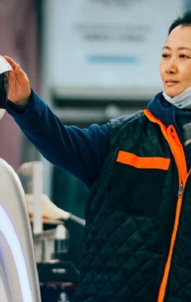 Na pierwszym planie azjatycka kobieta z jednorazową maską na twarzy, ubrana w bluzę i ortalionową obszerną kamizelę. Kobieta dotyka stojącego obok niej robota. Za jej plecami nieostry widok na wnętrze wyglądające na halę magazynową z ubraniami na wieszakach.