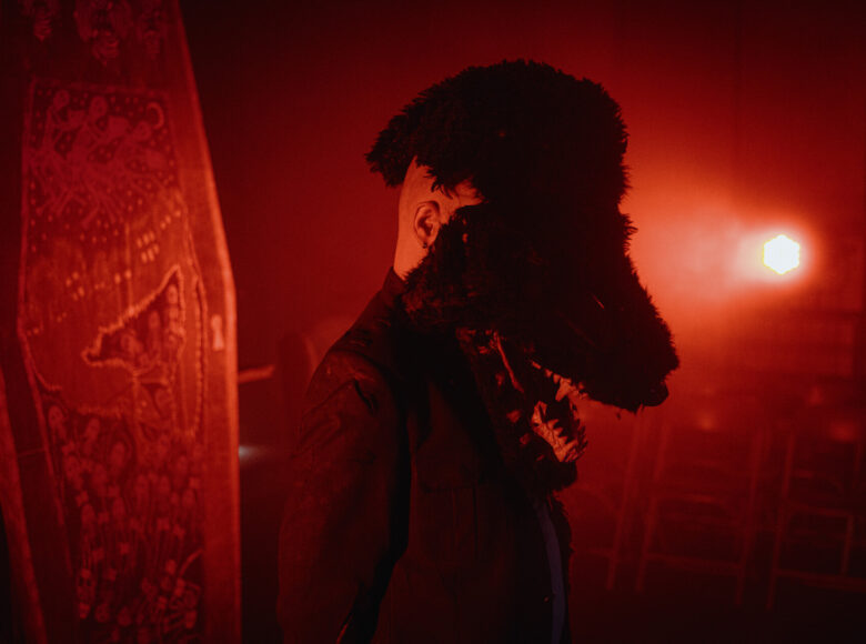 W pomieszczeniu oświetlonym czerwonym, ciemnym światłem stoi mężczyzna w kurtce. Na głowie ma maskę niedźwiedzia czy też wilka.