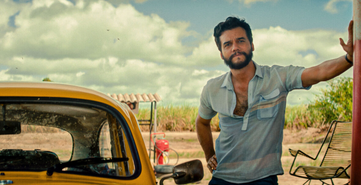 Na pierwszym planie młody, brodaty mężczyzna w rozpiętej koszulce polo. Mężczyzna stoi obok zaparkowanego samochodu. Za mężczyzną widoczny fragment ściany budynku oraz krzesło ogrodowe stojące przy budynku. Na dalszym planie gęste kępy trawy rosnące wzdłuż linii horyzontu oraz niebo pokryte białymi chmurami.