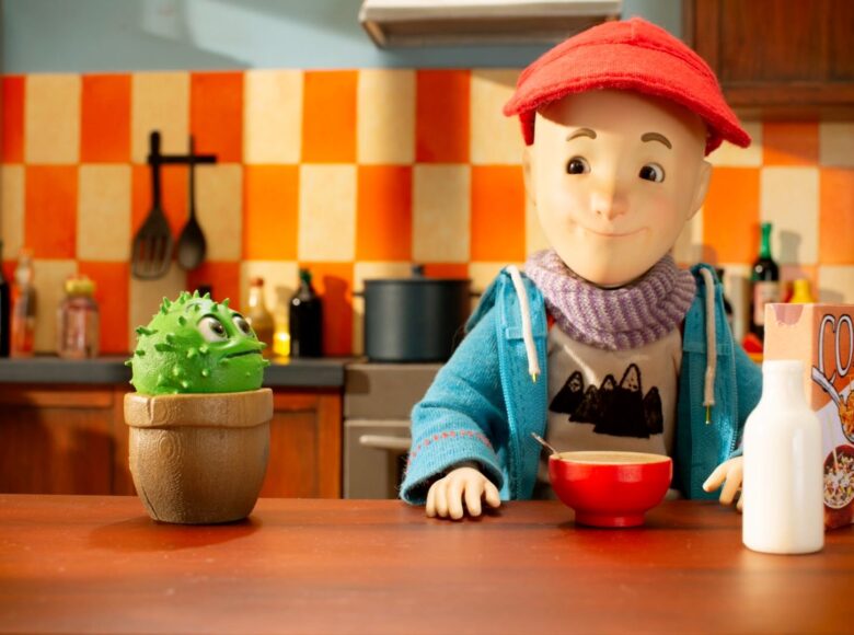 Kadr z filmu animowanego. W kuchni przy stole siedzi chłopiec ubrany w kurtkę, szalik i czapkę. Chłopcxyk spożywa płatki z mlekiem.