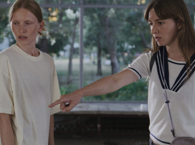 Obok siebie stoją dwie młode dziewczyny. Obie patrzą na coś poza kadrem, a jedna z nich wskazuje też palcem na to coś. W tle za dziewczynami duże okno, przez które widać zielone drzewa.