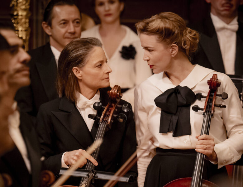 Filharmonia. Zbliżenie na grupę muzyczek i muzyków siedzących na scenie. Wszyscy ubrani są w eleganckie, czarno białe garnitury i kostiumy. Pośrodku siedzą obok siebie dwie kobiety z wiolonczelami. Zwrócone są do siebie twarzami.