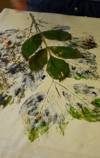 Zbliżenie na rozłożoną na stole tkaninę, na której namalowane są liście. Na tkaninie leży także prawdziwy liść.