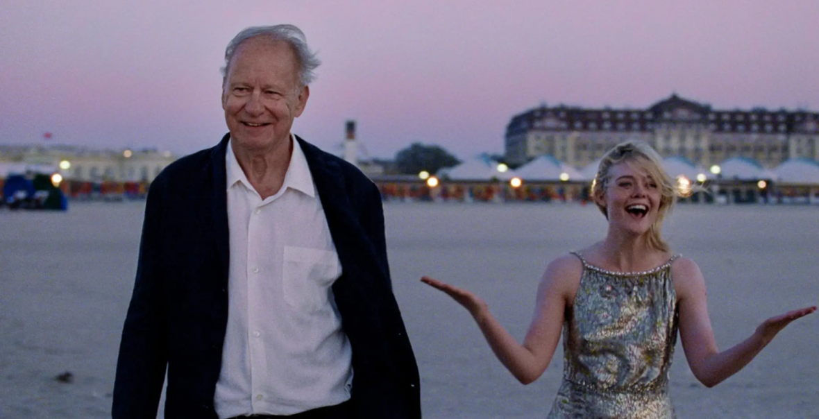 Zmierzch w mieście. Na pierwszym planie stoją obok siebie starszy mężczyzna i moda kobieta, oboje elegancko ubrani. Uśmiechają się. Za ich plecami widok na budynki i zapalające się światła.