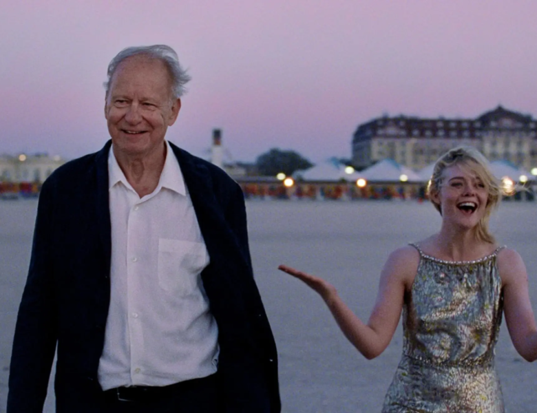 Zmierzch w mieście. Na pierwszym planie stoją obok siebie starszy mężczyzna i moda kobieta, oboje elegancko ubrani. Uśmiechają się. Za ich plecami widok na budynki i zapalające się światła.