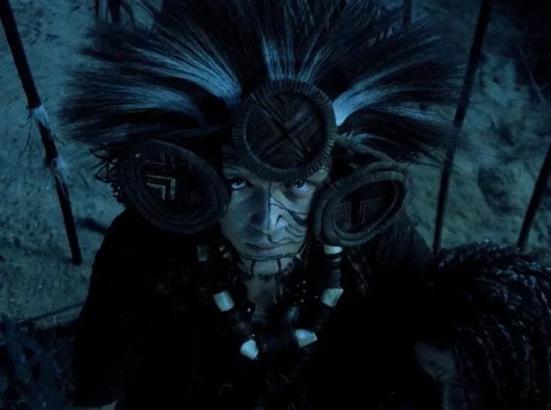 Zbliżenie na kobietę w ubraną w szamańskim stylu w szatę, ciężkie naszyjniki i strojne nakrycie głowy. Kobieta patrzy do góry. Obok niej widzimy drugą kobietę z włosami zaplecionymi w liczne warkoczyki.