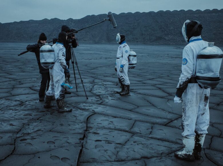 Na ziemi pokrytej pyłem węglowym stoi dwoje filmowców z kamerą oraz trzy osoby w kombinezonach ochronnych. Jedna z osób w kombinezonie wypowiada się do kamery. W tle wielka hałda węgla, a nad nią zasnute pyłem niebo.