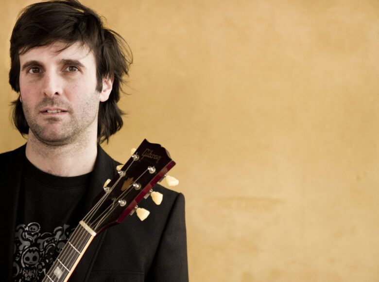 Do fotografii pozuje ciemnowłosy mężczyzna w średnim wieku, w czarnej bluzce i z gitarą. Za jego plecami żółta ściana.