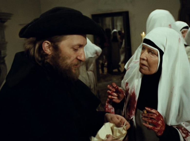 Scena historyczna. W dużym pomieszczeniu na pierwszym planie stoi mężczyzna w czarnym okryciu i czarnym, staroświeckim kapeluszu. Trzyma w dłoniach biały, niedookreślony przedmiot i przygląda mu się. Naprzeciwko niego stoi kobieta w stroju zakonnicy. Jej ręce i ubranie poplamione są czerwoną cieczą, która może kojarzyć się z krwią. Kobieta patrzy na mężczyznę. Na drugim planie stoi kilka kolejnych kobiet w strojach zakonnych, także poplamionych na czerwono. W głębi widzimy drzwi do innego pomieszczenia, w którym stoi kilka kolejnych osób.