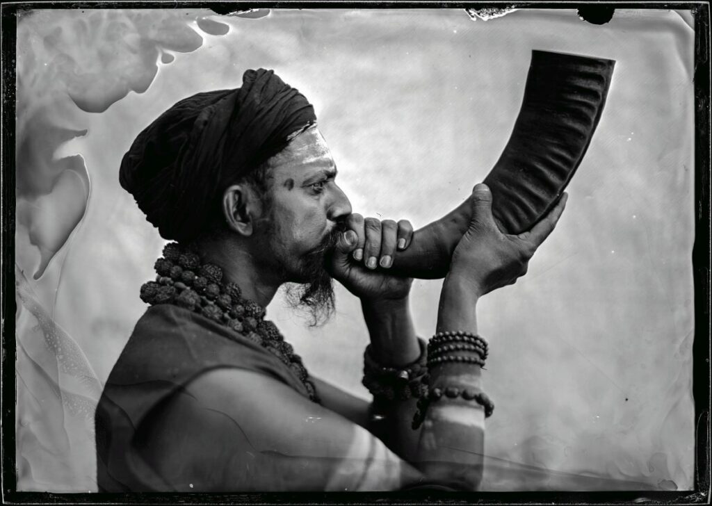 Czarno-biała fotografia przedstawia mężczyznę o hinduskich rysach twarzy i ciemnej karnacji, z wąsami i brodą. Ma na głowie ciemny turban i dmie w zakrzywiony róg. Ubrany jest w ciemną kamizelkę odsłaniającą nagie, umięśnione ramiona. Na szyi i przedramionach ma ozdoby przypominające sznury korali. Jego czoło jest pomarszczone, a twarz ma poważny, skupiony wyraz.