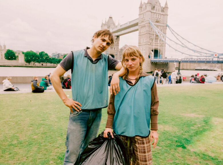 Miasto nad rzeką. Na trawniku nad brzegiem rzeki stoją dziewczyna z chłopakiem, pozując do zdjęcia. Dziewczyna trzyma w ręku wypchany worek na śmieci. Za nimi widać ludzi zgromadzonych na brzegu, pod zabytkowym mostem z wieżami. Na ostatnim planie, na drugim brzegu rzeki widnieje zabytkowa, reprezentacyjna budowla.