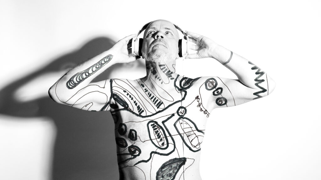 Czarno-biała fotografia przedstawia półnagiego mężczyznę w sile wieku, z krótkim siwym zarostem na twarzy o regularnych rysach i ogoloną głową. Jego ciało pokryte jest różnymi wzorami czarnej farby. Mężczyzna ma na uszach słuchawki, które podtrzymuje dłońmi na wysokości uszu. Stoi na białym tle, na które jego ciało rzuca cień po prawej stronie.
