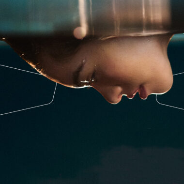 Na czarnym tle, u góry kadru widzimy fotografię kobiecej twarzy profilem. Twarz kobiety skierowana jest w dół.