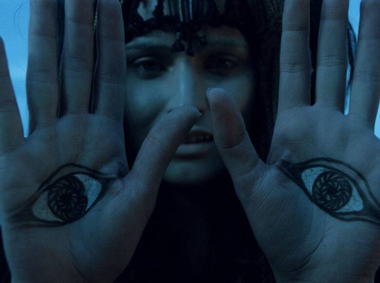 Zbliżenie na kobietę, która wyciąga w naszą stronę dłonie, Na każdej dłoni ma namalowane oko. Za kobietą niedookreślone, niebieskie tło.