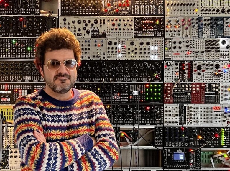 Do fotografii pozuje brodaty mężczyzna w średnim wieku, ubrany w kolorowy sweter i okulary przeciwsłoneczne. Za nim na ścianie półki zastawione ciasno jakimiś sprzętami elektronicznymi z pokrętadłami, przyciskami i kablami.