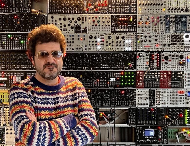 Do fotografii pozuje brodaty mężczyzna w średnim wieku, ubrany w kolorowy sweter i okulary przeciwsłoneczne. Za nim na ścianie półki zastawione ciasno jakimiś sprzętami elektronicznymi z pokrętadłami, przyciskami i kablami.