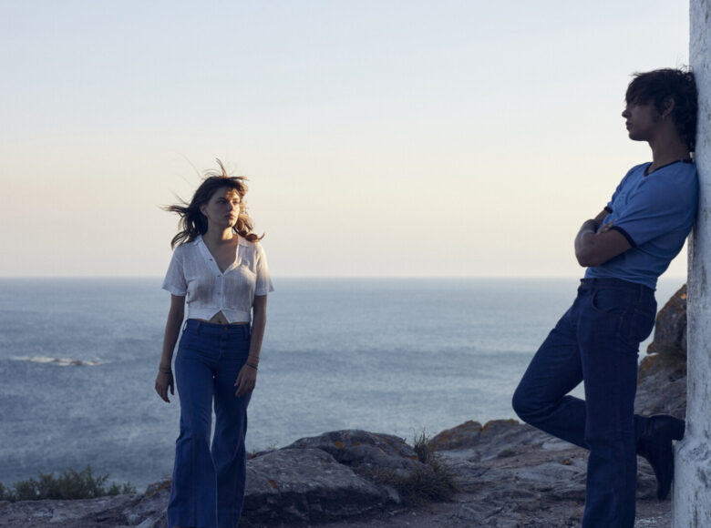 Schyłek dnia. Na skalistym brzegu morza stoi twarzą do nas młoda kobieta. Niedaleko niej profilem do nas stoi młody chłopak. Chłopak opiera się o ścianę jakiejś budowli, która w większej części znajduje się poza kadrem.