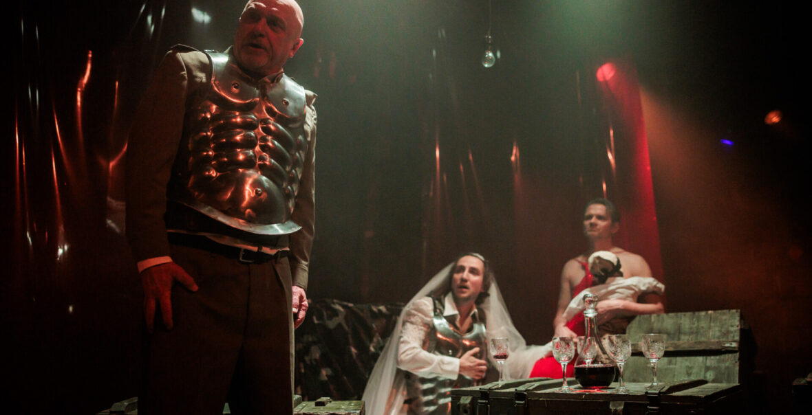 Spektakl teatralny. Na pierwszym planie mężczyzna w spodniach i w napierśniku, czyli fragmencie zbroi. Na dalszym planie dwóch mężczyzn siedzi przy drewnianej skrzyni zastawionej jak stół kieliszkami i karafką z czerwonym płynem. Jeden z mężczyzn ma na sobie suknię ślubną, welon i również napierśnik. Drugi ma czerwoną suknię i trzyma na rękach lakę wyglądającą jak niemowlę. Za nimi ciemne tło.