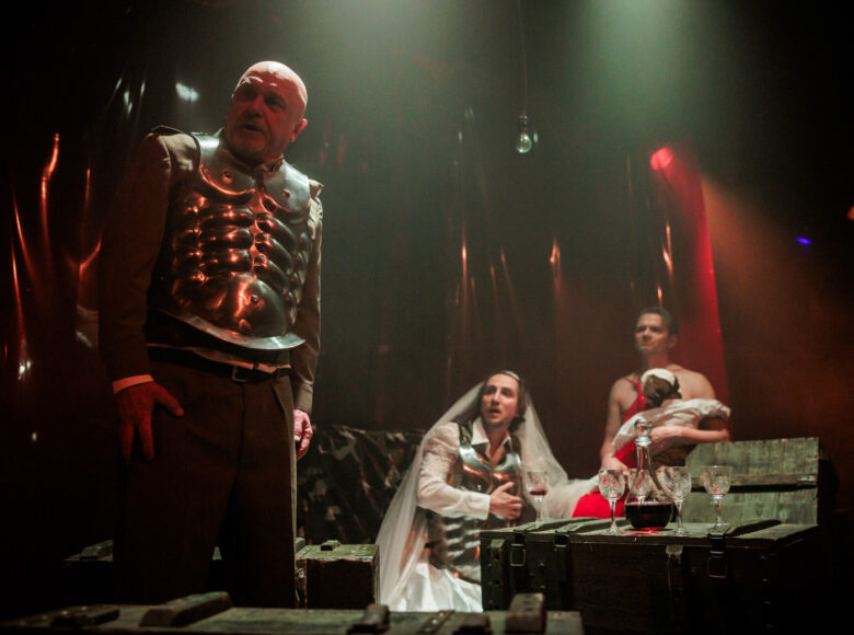 Spektakl teatralny. Na pierwszym planie mężczyzna w spodniach i w napierśniku, czyli fragmencie zbroi. Na dalszym planie dwóch mężczyzn siedzi przy drewnianej skrzyni zastawionej jak stół kieliszkami i karafką z czerwonym płynem. Jeden z mężczyzn ma na sobie suknię ślubną, welon i również napierśnik. Drugi ma czerwoną suknię i trzyma na rękach lakę wyglądającą jak niemowlę. Za nimi ciemne tło.