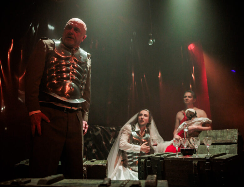 Spektakl teatralny. Na pierwszym planie mężczyzna w spodniach i w napierśniku, czyli fragmencie zbroi. Na dalszym planie dwóch mężczyzn siedzi przy drewnianej skrzyni zastawionej jak stół kieliszkami i karafką z czerwonym płynem. Jeden z mężczyzn ma na sobie suknię ślubną, welon i również napierśnik. Drugi ma czerwoną suknię i trzyma na rękach lakę wyglądającą jak niemowlę. Za nimi ciemne tło.