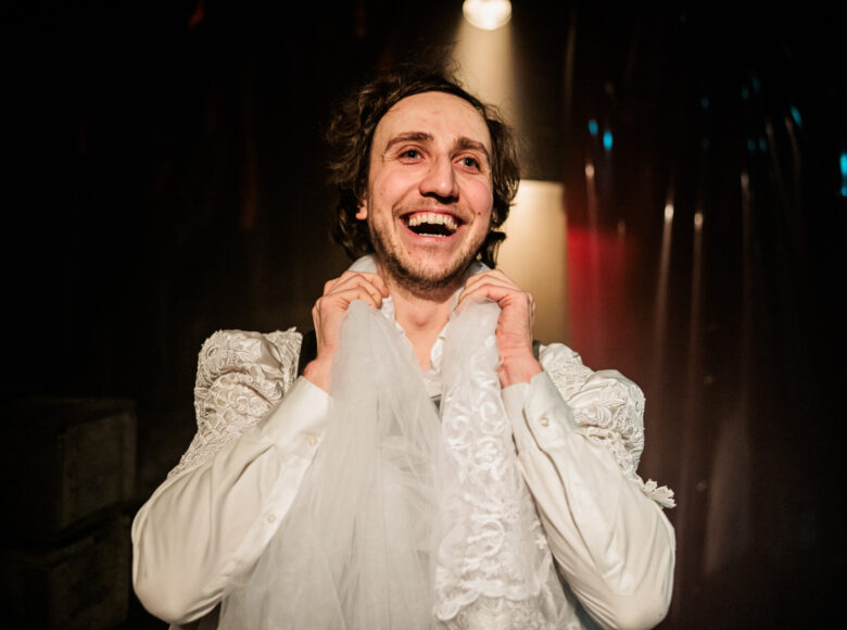 Zbliżenie na młodego, roześmianego mężczyznę. Mężczyzna ubrany jest w damską bluzkę z bufkami, w białym kolorze. Za mężczyzną czarne tło i jasny punkt światła z reflektora.