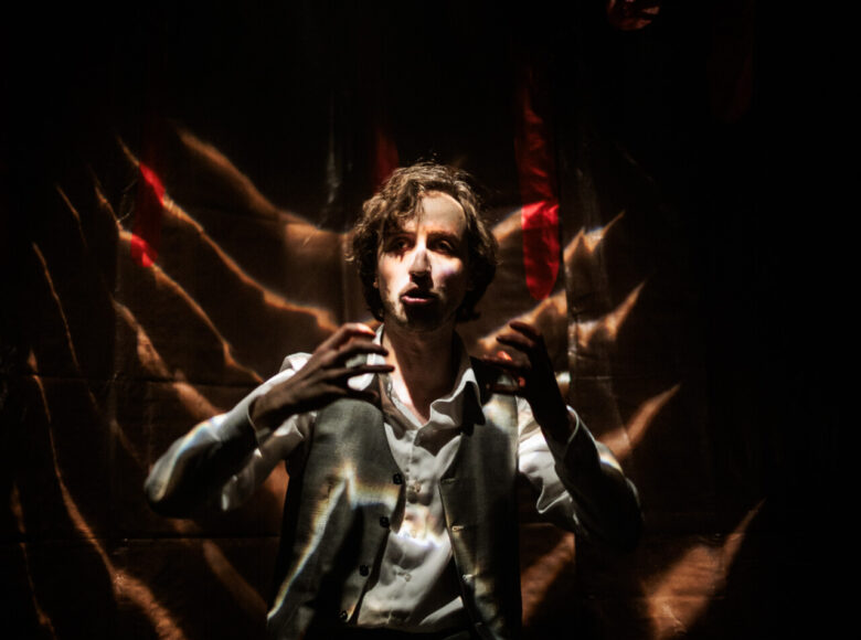 Spektakl teatralny. Na fotelu oświetlonym punktowymi światłami siedzi mężczyzna. Przestrzeń dookoła pogrążona jest w ciemności. Mężczyzna unosi ręce do góry i coś mówi. Ubrany jest w spodnie, koszulę i kamizelkę.