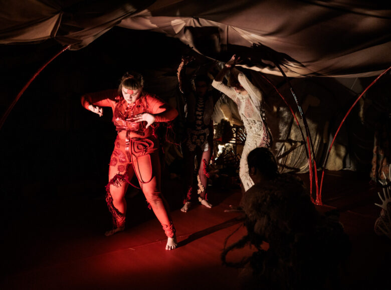 Spektakl teatralny. Pod rozpiętą niczym namiot tkaniną stoją trzy kobiety w dynamicznych pozach. Ubrane są w obcisłe kombinezony ozdobione koronkami i różnorodnymi naszyciami. Jedna z kobiet ma kombinezon czerwony, druga biały, trzecia czerwono-biało-czarny.
