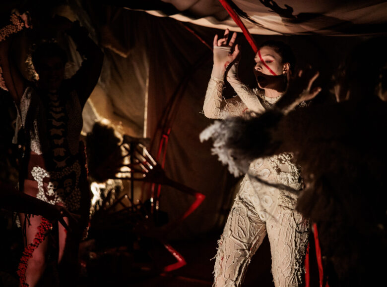 Spektakl teatralny. Pod rozpiętą jak namiot tkaniną stoją obok siebie dwie kobiety. Obie unoszą ręce do góry. Są ubrane w obcisłe kombinezony, ozdobione koronkami i naszyciami. Jedna ma kombinezon biały, druga biało czarno czerwony.