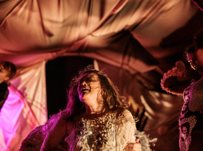 Zbliżenie na kobietę ubraną na biało. Nad nią rozciągnięta jest tkanina jak namiot.