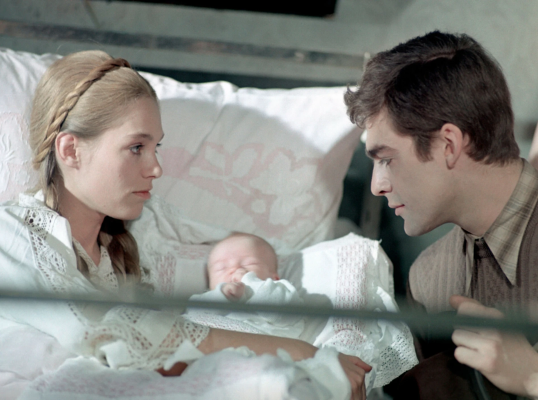 Młoda kobieta siedzi w łóżku, trzymając na rękach śpiące niemowlę. Naprzeciwko kobiety, przy łóżku siedzi młody mężczyzna. Mężczyzna patrzy na dziecko, kobieta patrzy na mężczyznę.