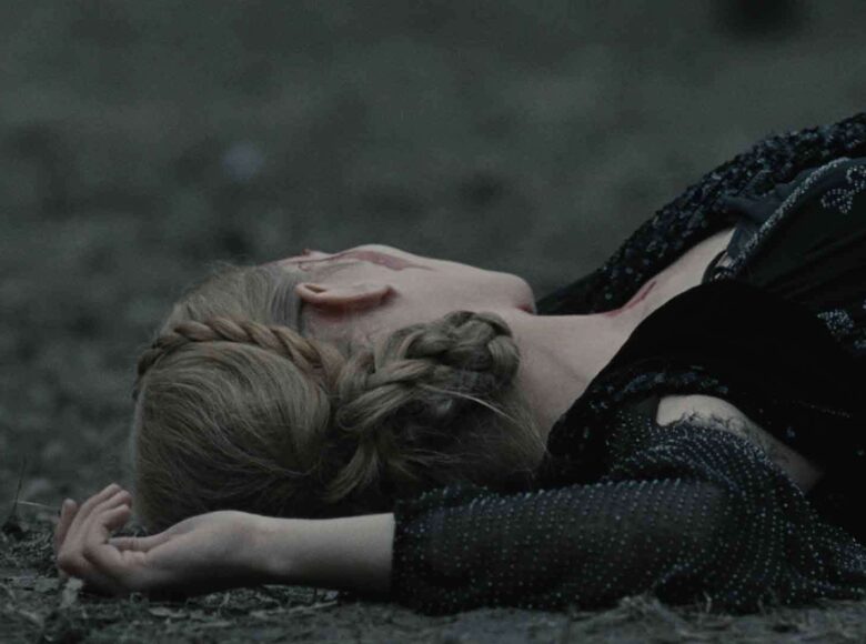 Zbliżenie na głowę i ramiona dziewczyny, leżącej na ziemi, pokrytej kawałkami uschniętych gałązek czy łodyg. Dziewczyna odwraca głowę od nas w drugą stronę. Uczesana jest w warkocze spięte na głowie, ubrana jest w elegancką sukienkę i narzutkę.