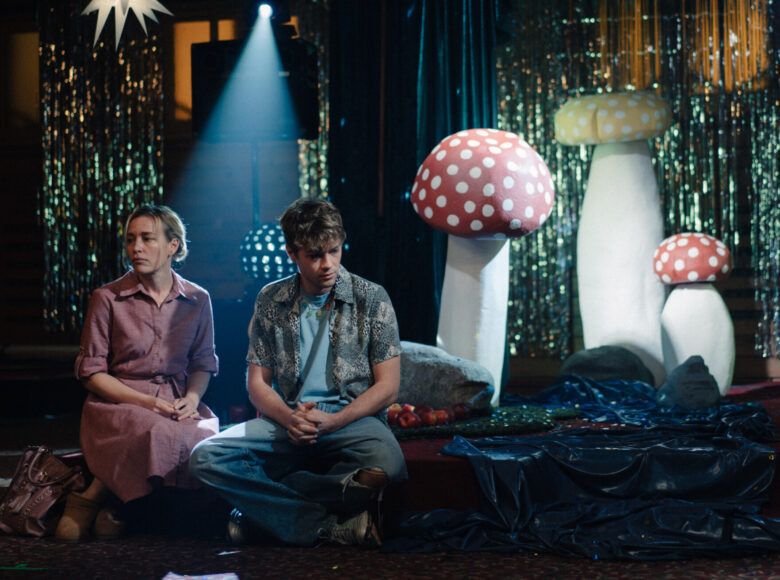 Niedookreślona przestrzeń, która może kojarzyć się ze scenografią teatralną. Półmrok. Niski podest, na którym siedzą obok siebie kobieta z młodym mężczyzną. Za nimi stoją elementy dekoracyjne, przedstawiające grzyby nadnaturalnej wielkości. W tle wiszą zasłony wykonane z błyszczących tasiemek i świeci punktowo reflektor.