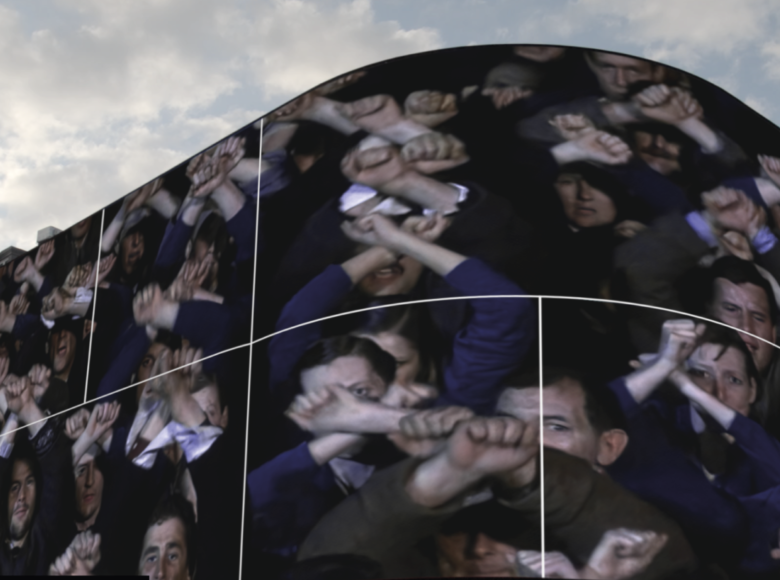 Ciąg zabytkowych kamienic wzdłuż ulicy. Na rogu jednej z kamienic zawieszony wielki ekran. Na ekranie widzimy zbliżenie na twarze stojących obok siebie ludzi. Unoszą oni do góry ręce zaciśnięte w pięści.