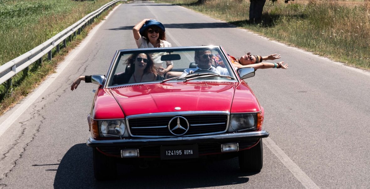 Lato. Wąska szosa. Szosą w naszą stronę jedzie czerwony kabriolet. W kabriolecie podróżują cztery dorosłe osoby płci obojga, wszyscy w okularach słonecznych. Jedna z kobiet wychyla się do tyłu, druga z uśmiechem przytrzymuje swój kapelusz. Po obu stronach szosy rośnie trawa.