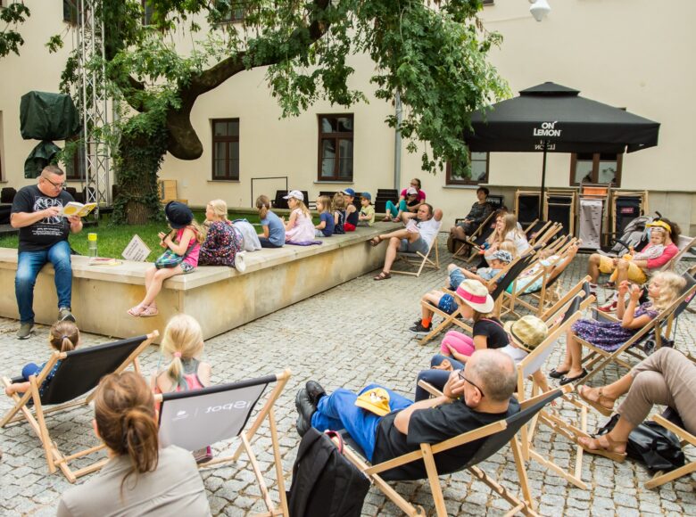 Wirydarz Centrum Kultury w Lublinie. na murku siedzi aktor Dariusz Jeż i czyta na głos książkę trzymaną przez siebie w ręku. Wokół niego, na murku i na leżakach rozstawionych na ziemi, siedzą słuchający go ludzie - dzieci i dorośli.