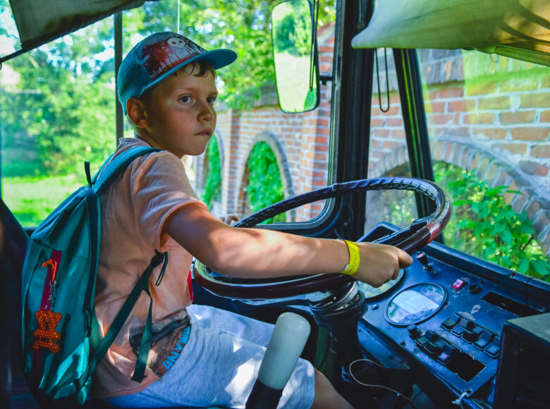 Wnętrze kabiny kierowcy w samochodzie ciężarowym lub też w autobusie. Pojazd ten jest zaparkowany przed ceglastym murem. Za kierownicą siedzi kilkuletni chłopiec.