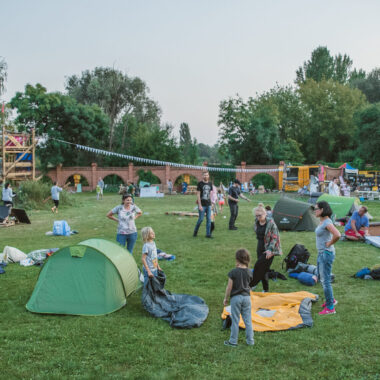 W plenerze, na trawie rozbite namioty. Wokół namiotów chodzą ludzie. W tle zielone drzewa.